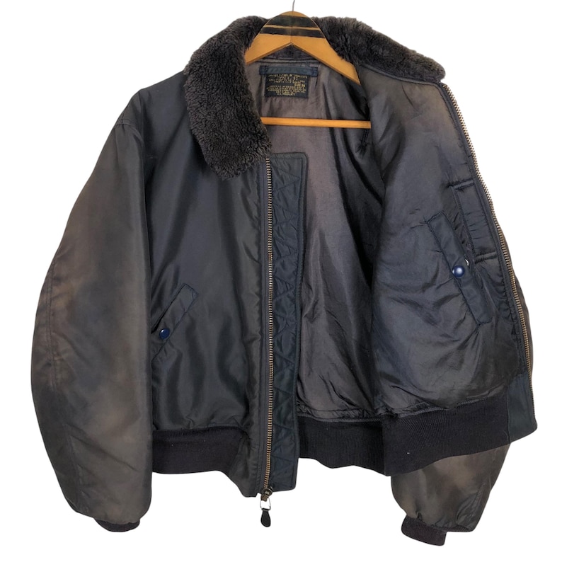 May include: A vintage black bomber jacket with a faux fur collar. The jacket features a zipper closure, multiple pockets, and ribbed cuffs and hem. The jacket is hanging on a wooden hanger.