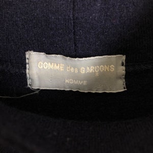 May include: Close-up of a navy blue garment with a label. The label is rectangular, with the words "COMME des GARCONS" in gold lettering at the top and "HOMME" below. The label is attached to the garment.