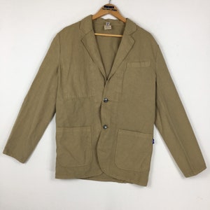 Puede incluir: Un blazer color beige con dos bolsillos delanteros, un bolsillo en el pecho y dos botones grises oscuros. La chaqueta tiene mangas largas y solapas con muescas. Una pequeña etiqueta es visible en la percha de madera.
