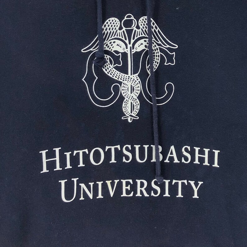 May include: Navy blue hooded sweatshirt featuring a white caduceus emblem and the text "Hitotsubashi University". The hoodie has a drawstring hood and is made of a soft material. The text is in a serif font.