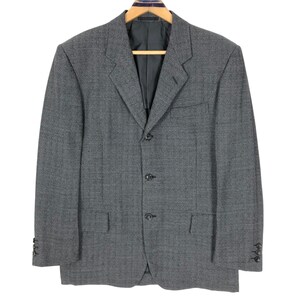 Puede incluir: Un blazer gris con cierre de tres botones, dos bolsillos delanteros y puños abotonados. El blazer está colgado de una percha de madera. La tela tiene un sutil patrón texturizado.