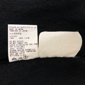 May include: A black fabric with two white tags. One tag has text in English and French, including "MADE IN JAPAN" and care instructions. The other tag is a blank, white, curved shape.