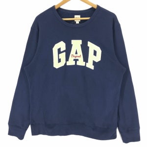 May include: Navy blue GAP sweatshirt with a crew neck. The word "GAP" is printed in large, off-white block letters across the chest, with the word "Original" in red script underneath. The sweatshirt is hanging on a wooden hanger.