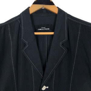 May include: A black blazer with white stitching details. The blazer features a notched lapel, a single-button closure, and the brand name "tricot COMME des GAR&Ccedil;ONS" on a label. The jacket is hanging on a wooden hanger.