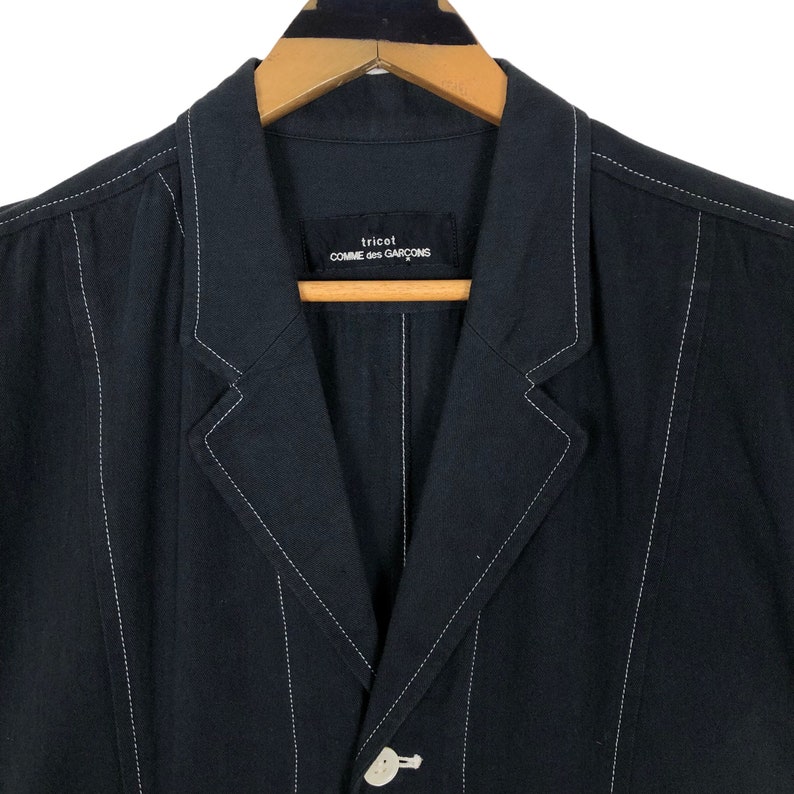 May include: A black blazer with white stitching details. The blazer features a notched lapel, a single-button closure, and the brand name "tricot COMME des GAR&Ccedil;ONS" on a label. The jacket is hanging on a wooden hanger.