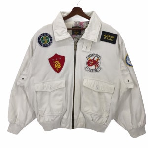 Könnte beinhalten: Weiße Bomberjacke mit mehreren Aufnähern, darunter "SCORPIONS" und "AVIATOR U.S.A.F." Aufnäher. Sie hat zwei Vordertaschen und einen Reißverschluss. Die Jacke hat einen gerippten Kragen und Bündchen. Das Innenfutter hat ein Blumenmuster.