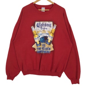 Op de afbeelding: Rode sweatshirt met een Corona Extra-afbeelding. De afbeelding toont twee bierflessen, het Corona Extra-logo en de tekst "IT'S FIVE O'CLOCK SOMEWHERE!". De sweatshirt heeft lange mouwen en een ronde hals.