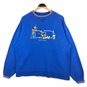 Puede incluir: Sudadera azul con un diseño de Mickey Mouse, Donald Duck, Goofy y Pluto. El diseño incluye la letra "D" con los personajes y el texto "EST. 1928". El cuello y los puños tienen rayas rojas, blancas y azules.