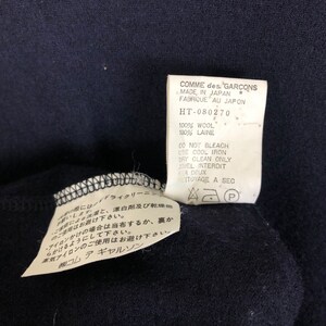 May include: Close-up of a navy blue garment with two attached tags. The tags display text including "COMME des GARCONS MADE IN JAPAN" and care instructions. The garment is made of 100% wool.