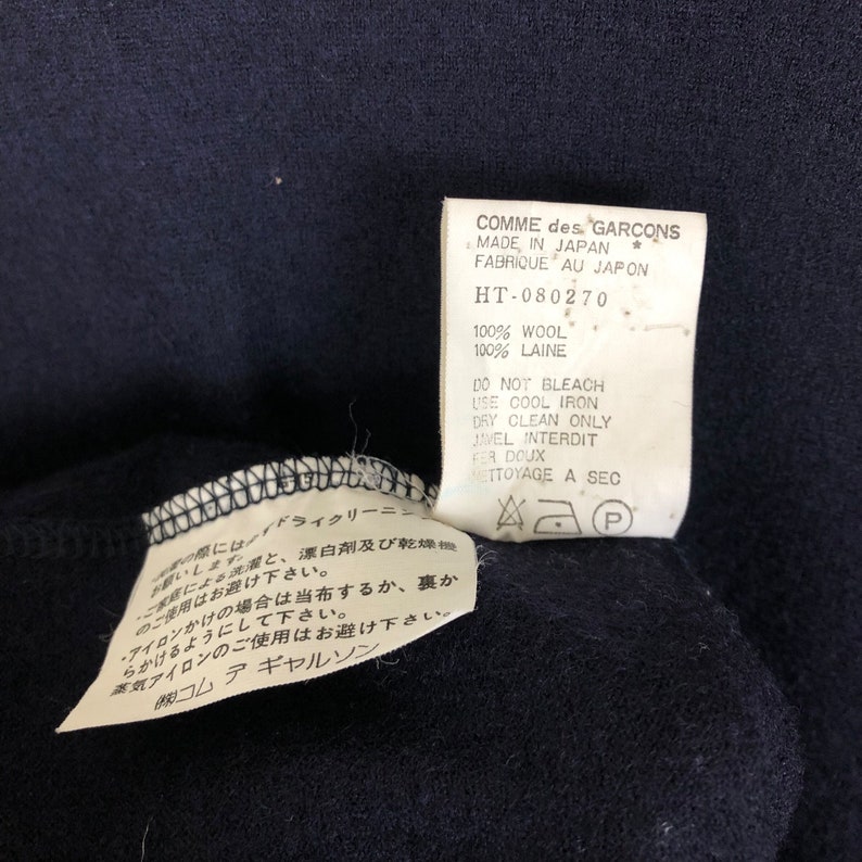 May include: Close-up of a navy blue garment with two attached tags. The tags display text including "COMME des GARCONS MADE IN JAPAN" and care instructions. The garment is made of 100% wool.