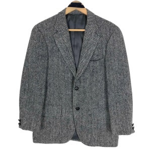 May include: A grey herringbone blazer with a classic three-button fastening. The jacket has two front pockets, a chest pocket, and buttoned cuffs. It is hanging on a wooden hanger against a white background.