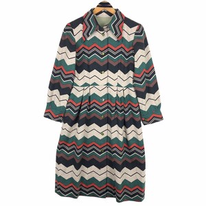 Może przedstawiać: Sukienka z długim rękawem z kolorowym wzorem w zygzaki w kolorze zielonym, czerwonym, czarnym i beżowym. Sukienka ma kołnierz, zapinana na guziki z przodu i plisowaną spódnicę. Sukienka wisi na drewnianym wieszaku.