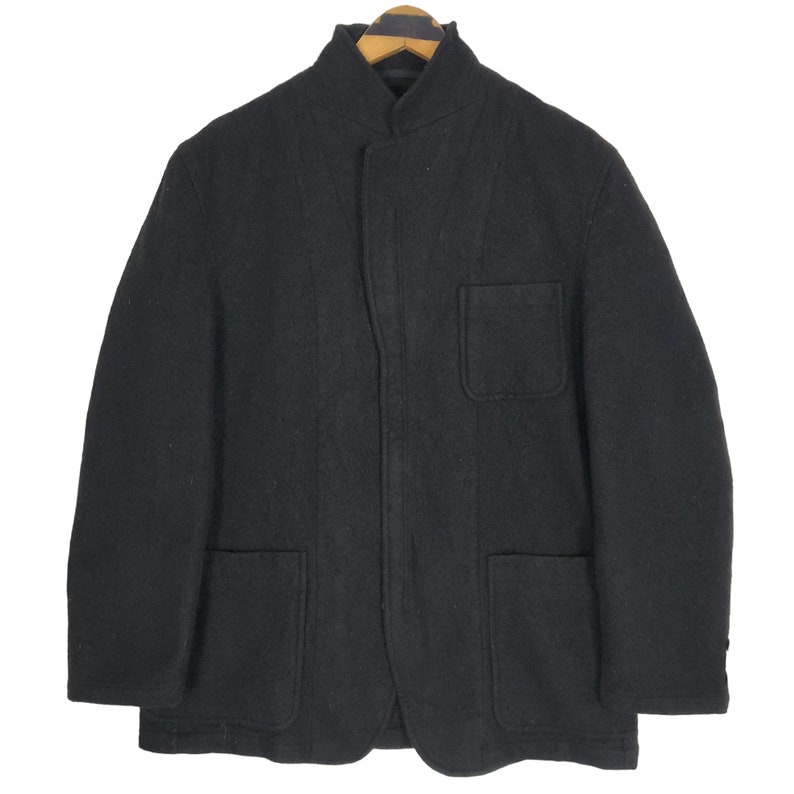 May include: A black wool jacket with a mandarin collar and two front pockets. The jacket is hanging on a wooden hanger. The jacket has a simple, minimalist design.