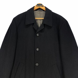 May include: A black wool coat with a classic design. The coat features a notched collar, three-button closure, and two front pockets. The coat is hanging on a wooden hanger. The interior lining is a light gray color.