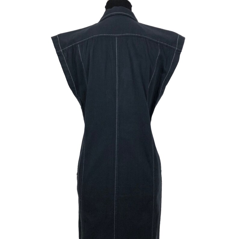 May include: A sleeveless black dress with white stitching details. The dress has a collared neckline and a button-down front. The white stitching outlines the seams and edges of the dress, creating a contrasting design. The dress is displayed on a mannequin.
