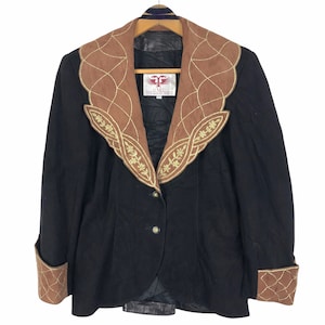 Pode incluir: Blazer preto com gola e punhos em camurça castanha, com bordados dourados. A gola e os punhos apresentam um design geométrico. O blazer tem fecho de dois botões e uma etiqueta que diz "G.M.P. ESTA MARINO DESIGN".