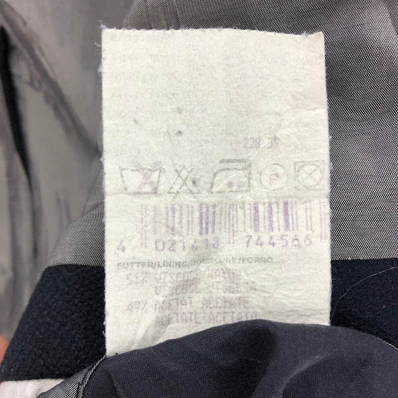 May include: A close-up of a clothing tag with a barcode and various symbols. The tag is white with black text and a purple barcode. The tag is attached to a dark garment.