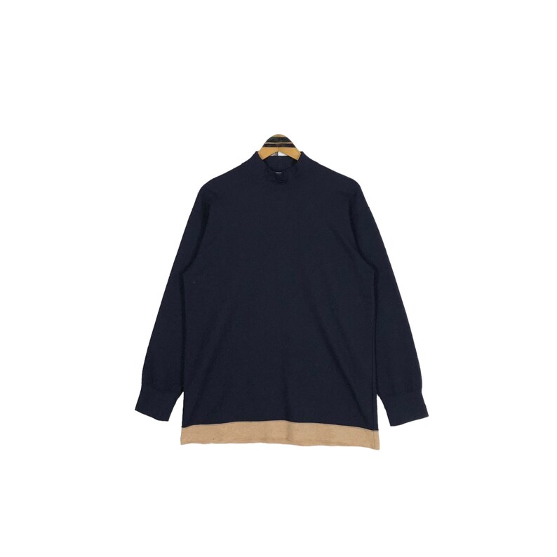 May include: A navy blue long-sleeve shirt with a mock neck and a tan-colored bottom hem. The shirt is hanging on a wooden hanger against a white background. The shirt is a solid color.