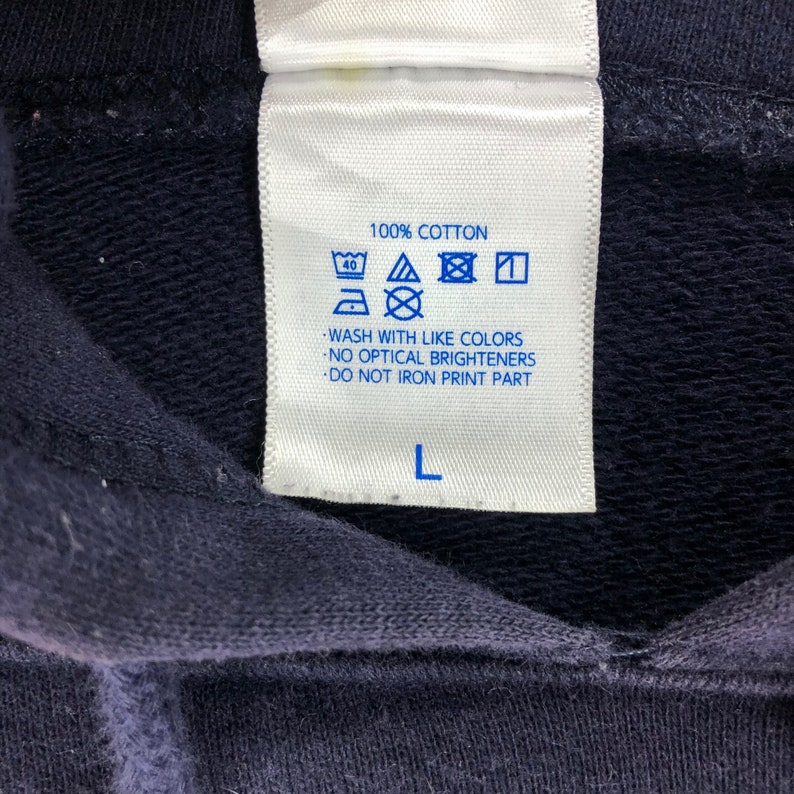 May include: Close-up of a navy blue garment tag with care instructions. The tag reads "100% COTTON" and includes washing symbols. The text "WASH WITH LIKE COLORS" and "DO NOT IRON PRINT PART" are also visible. The size is "L".