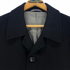 May include: Close-up of a black coat with a notched collar and a single button closure. The coat is hanging on a wooden hanger. The interior lining is a light gray color.