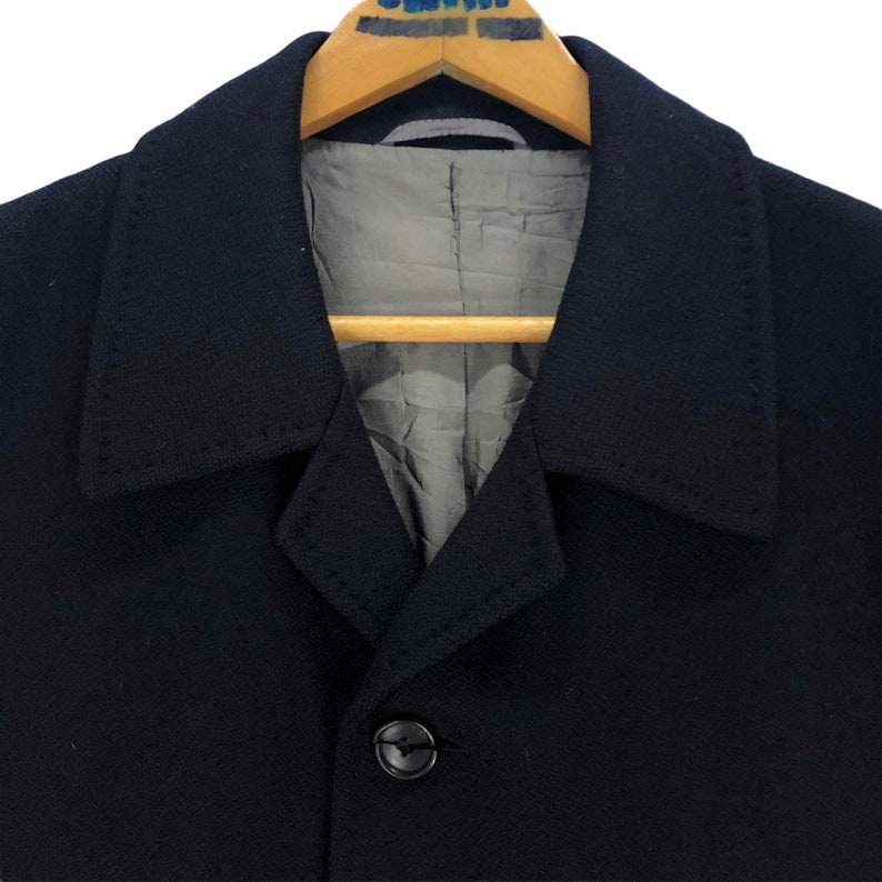 May include: Close-up of a black coat with a notched collar and a single button closure. The coat is hanging on a wooden hanger. The interior lining is a light gray color.