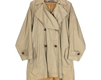 Op de afbeelding: Een beige trenchcoat met een dubbele rij knopen, grote revers en knoopsluitingen. De jas, hangend aan een houten kleerhanger, heeft een bijpassende riem en zakken, wat een klassieke, op maat gemaakte stijl laat zien.