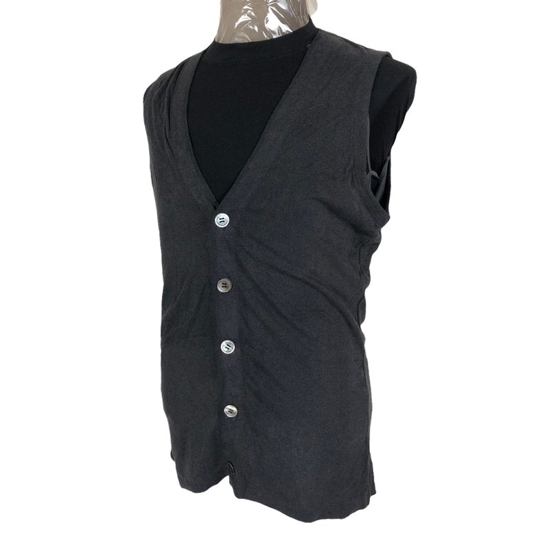May include: Dark gray vest with a V-neck and four round, silver-colored buttons. The vest is sleeveless and appears to be made of a soft, textured fabric. The vest is displayed on a mannequin.