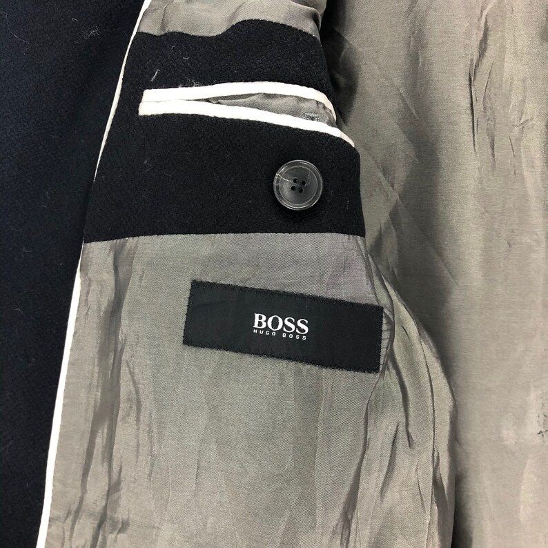 May include: Close-up of the interior of a black wool coat. The coat features a black lapel with a single button and a gray lining. A black label with the white text "BOSS HUGO BOSS" is sewn into the lining.
