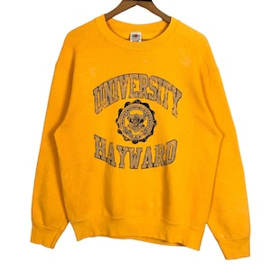 Könnte beinhalten: Senfgelbes Sweatshirt mit dem Text "UNIVERSITY HAYWARD" in einem verwaschenen Vintage-Stil. Das Sweatshirt hat einen Rundhalsausschnitt und lange Ärmel. Die Grafik enthält ein rundes Emblem mit zusätzlichem Text.