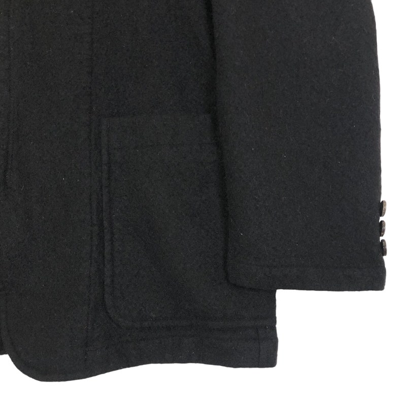 May include: Close-up of a black wool blazer. The blazer features a single pocket on the left side and three buttons on the sleeve. The fabric has a textured appearance.