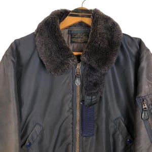 May include: A dark blue bomber jacket with a brown faux fur collar. The jacket has a zipper closure, a blue fabric detail, and a pocket with a zipper on the sleeve. The jacket is hanging on a wooden hanger.