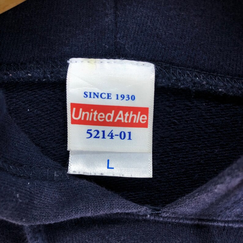 May include: Close-up of a navy blue hoodie label. The label is white with red and black text that reads "United Athle" and "5214-01". The label also includes the text "SINCE 1930" and the size "L".