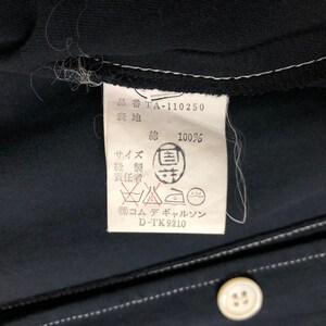 May include: Close-up of a black garment tag with white text and a white border. The tag includes Japanese characters, washing instructions, and the text "綿 100%" indicating 100% cotton. A white button is visible.