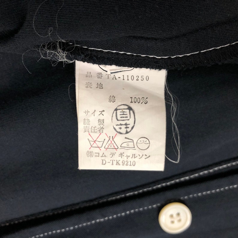 May include: Close-up of a black garment tag with white text and a white border. The tag includes Japanese characters, washing instructions, and the text "綿 100%" indicating 100% cotton. A white button is visible.