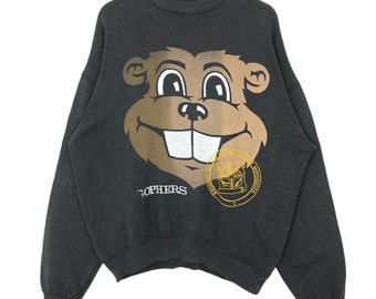 May include: Dark gray sweatshirt featuring a large cartoon gopher face in shades of brown and white. The sweatshirt has the word "Gophers" printed below the gopher face and a university seal. The sweatshirt is hanging on a wooden hanger.
