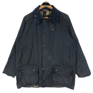 Puede incluir: Chaqueta azul marino oscuro con cuello de pana y cuatro bolsillos delanteros. La chaqueta tiene cierre de botones y forro a cuadros visible en el cuello y el dobladillo. La marca "Barbour" es visible en uno de los bolsillos.