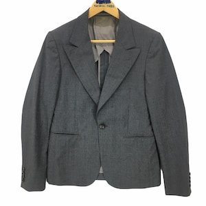 Puede incluir: Una blazer gris con cierre de un solo botón, solapas con muescas y un corte entallado. La chaqueta tiene tres botones en cada puño y un bolsillo en el pecho. Está colgada de una percha de madera.