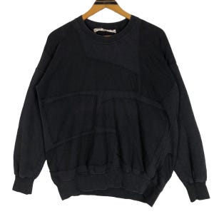 Puede incluir: Sudadera negra con un diseño de patchwork único. La parte superior de manga larga presenta cuello redondo y puños acanalados. La prenda tiene un corte cuadrado y un patrón de costura distintivo en la parte delantera.