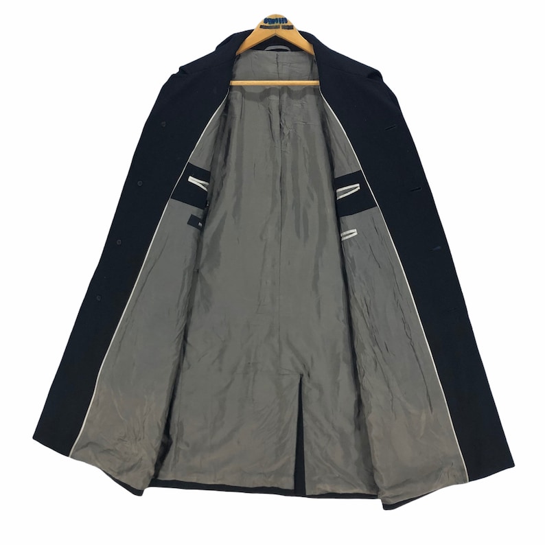 May include: A dark navy blue overcoat with a classic design. The coat is open, revealing a gray interior lining and white trim along the edges. The coat is hanging on a wooden hanger.
