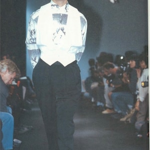 May include: A model on a runway wearing a white vest over a patterned shirt, black pants, and black shoes with white socks. The shirt has a black and white design. The model is standing in front of an audience.