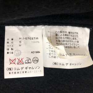 May include: Close-up of a black garment tag with Japanese text and care symbols. The tag includes the product code "HJ-07037M", material composition "毛 100%", and size "M". The tag also features the brand name and address.