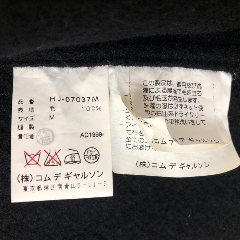 May include: Close-up of a black garment tag with Japanese text and care symbols. The tag includes the product code "HJ-07037M", material composition "毛 100%", and size "M". The tag also features the brand name and address.