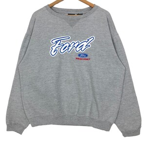 May include: A grey sweatshirt featuring the word "Ford" in blue script. Below the word "Ford" is the word "Racing" in red. The sweatshirt has a crew neck and long sleeves.