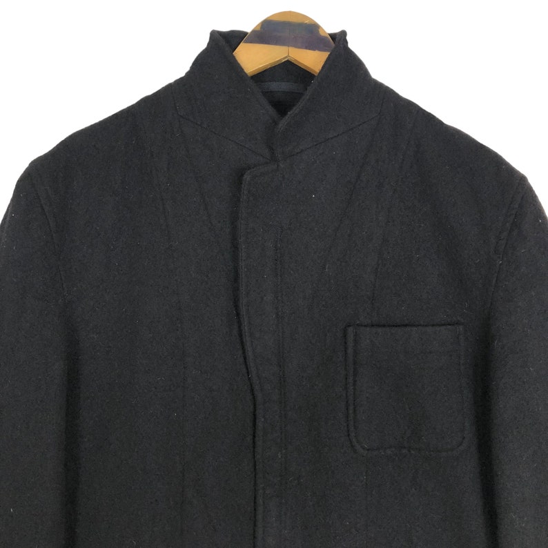 May include: A black wool coat with a mandarin collar and a single pocket. The coat is hanging on a wooden hanger. The coat has a minimalist design.