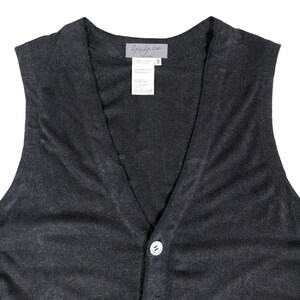 May include: A dark gray, sleeveless vest with a V-neck and a single white button closure. The vest has a textured appearance and a label at the back of the neck with the brand name and size information. The vest is made of a soft, possibly knit, material.