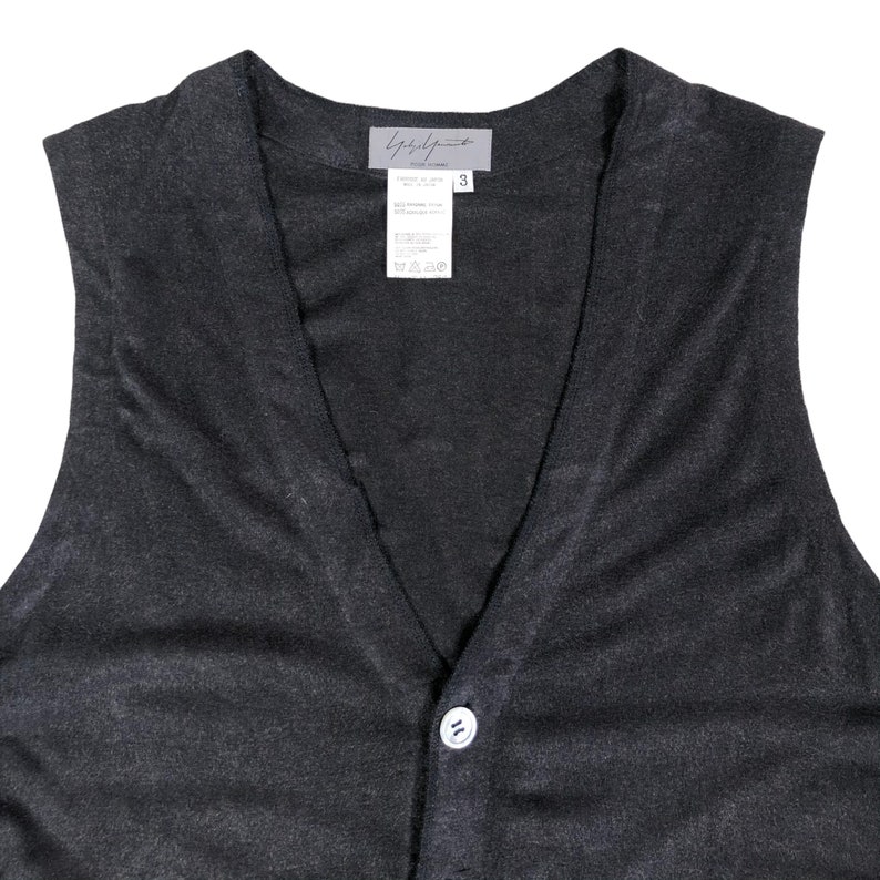 May include: A dark gray, sleeveless vest with a V-neck and a single white button closure. The vest has a textured appearance and a label at the back of the neck with the brand name and size information. The vest is made of a soft, possibly knit, material.