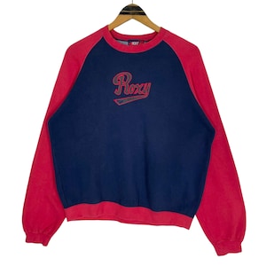 May include: A navy blue and red sweatshirt featuring the Roxy logo. The sleeves and upper shoulders are red, contrasting with the navy blue body. The Roxy logo is embroidered in a cursive style with a red outline.