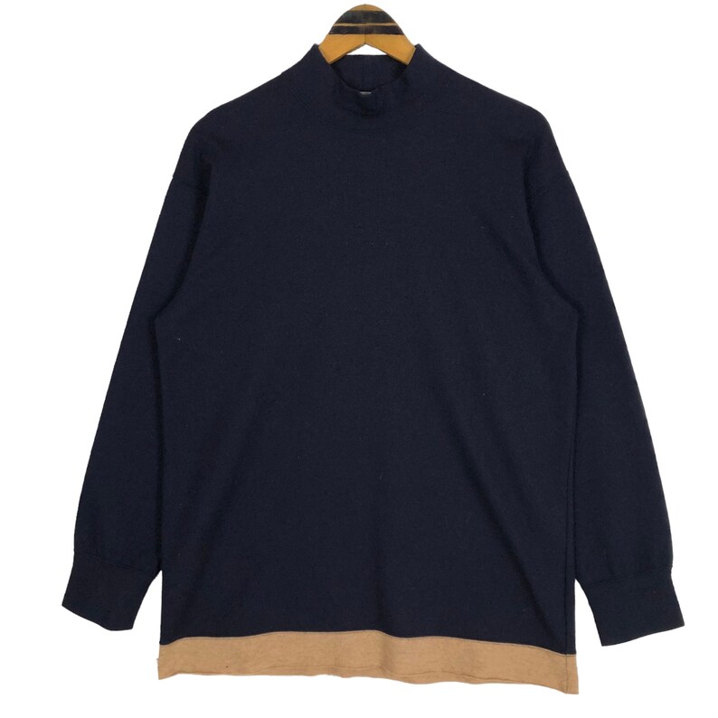 May include: Navy blue long-sleeve shirt with a mock neck and a tan-colored bottom hem. The shirt is hanging on a wooden hanger against a white background. The shirt's design is simple and classic.