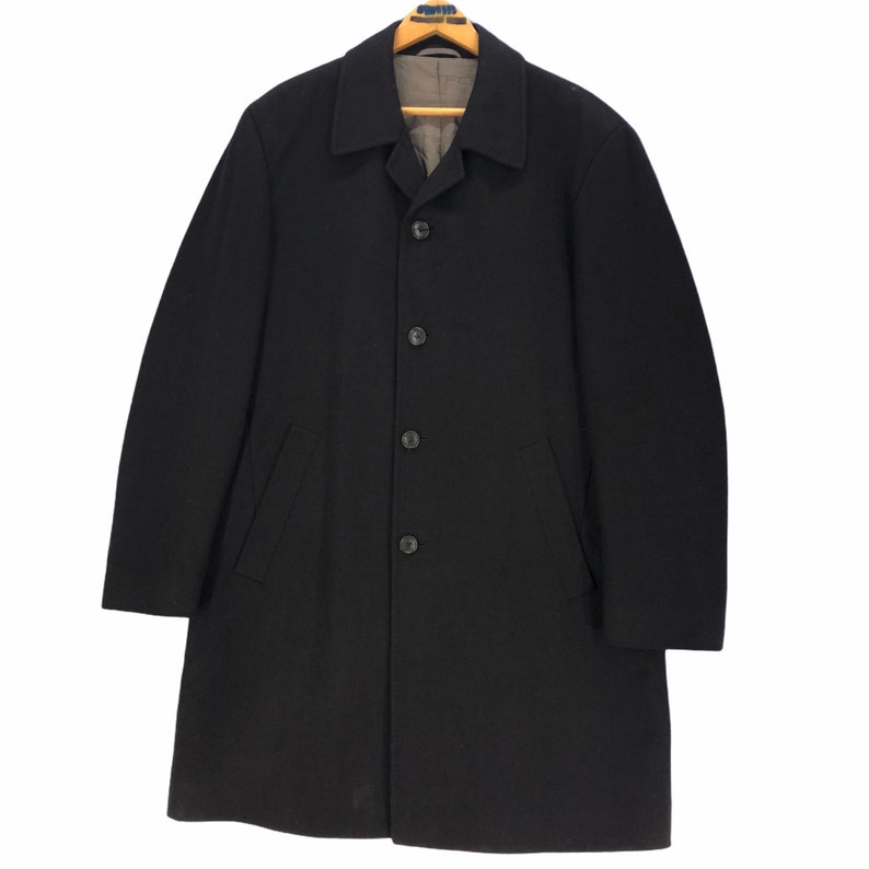May include: A black overcoat with a notched collar, four buttons, and two front pockets. The coat is hanging on a wooden hanger. The lining is a light gray color.