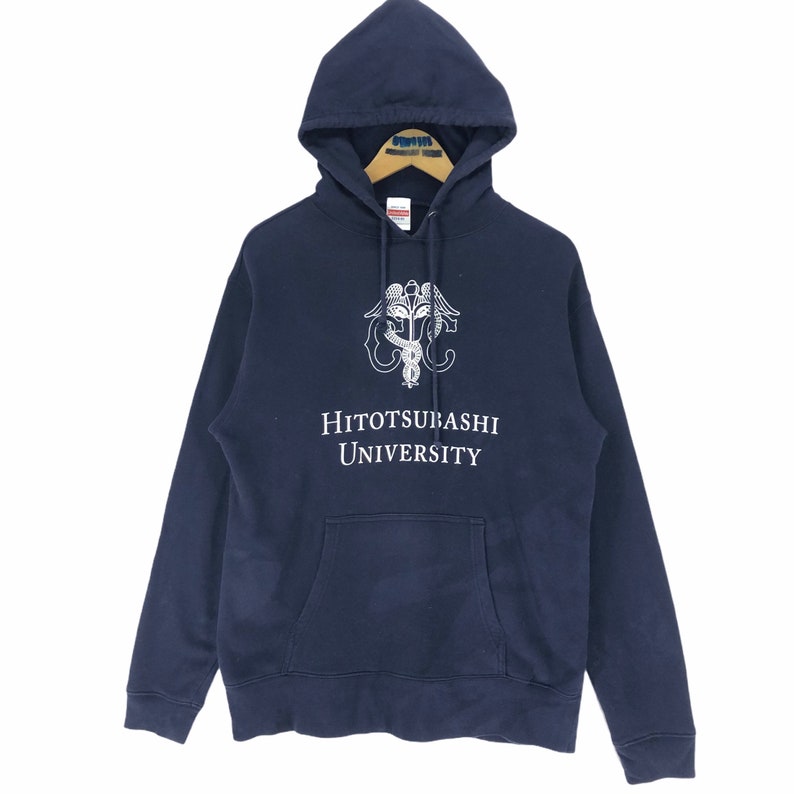 May include: Navy blue hooded sweatshirt with a front pocket. It features a white graphic and the text "Hitotsubashi University". The garment is displayed on a wooden hanger.